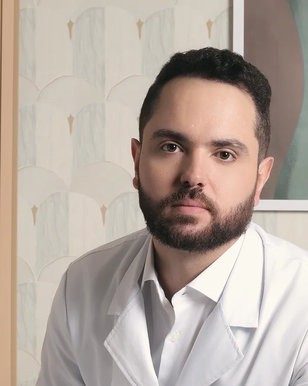 Retrato do Dr. Igor Reggiani Gomes, de jaleco branco, sentado à mesa, olhando para a câmera